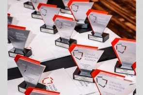 Kilka eleganckich, nowoczesnych statuetek o geometrycznych kształtach ustawionych na stole. Przeważają kolory srebrny, czarny i czerwony. Obok leżą białe teczki z logotypem organizatora. Nagrody są oznaczone nazwami miast i powiatów, które je otrzymały.