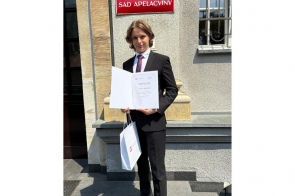 Młody chłopak stoi przed budynkiem i prezentuje dyplom oraz trzyma białą torbę. Na elewacji budynku widoczna jest tablica z napisem „Sąd Apelacyjny".