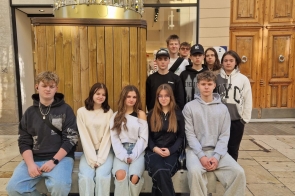 Dziewięć młodych osób ustawionych w dwóch rzędach – część siedzi na ławce, pozostali stoją za nimi. Ubrani w codzienne stroje w stonowanych kolorach. W tle drewniana zabudowa, duże drzwi z ozdobnymi uchwytami oraz przeszklone wejście do budynku.