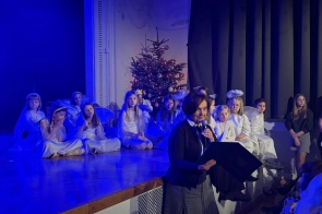 Janina Żagan Starosta Cieszyński przemawia do mikrofonu przy scenie, trzymając teczkę. Na scenie siedzą dzieci w białych strojach, a w tle widać choinkę i niebieskie oświetlenie.