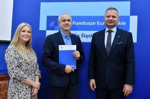 Trzy osoby stoją przed niebieską ścianką z napisem „Fundusze Europejskie dla Śląskiego”. Mężczyzna w środku trzyma w ręku niebieską teczkę z logo. Wszyscy ubrani są w stroje formalne – dwaj mężczyźni w garniturach, kobieta w jasnej wzorzystej sukience – i pozują do zdjęcia.