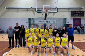 Drużyna koszykarska dziewcząt ustawiona do wspólnego zdjęcia na hali sportowej, w żółtych strojach, z trenerami i sztabem w tle oraz koszem do gry i tablicą wyników nad nimi.