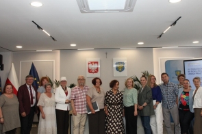 Grupa uczestników spotkania pozuje do wspólnego zdjęcia w sali sesyjnej. Osoby stoją w jednym rzędzie, część z nich trzyma dokumenty. 