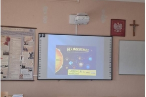 Zdjęcie przedstawia salę lekcyjną. Na ścianie wyświetlony jest slajd z prezentacji „Dermokosmos”. Przed ścianą ustawione są ławki i krzesła. Po lewej stronie widoczne plakaty edukacyjne, po prawej tablica.