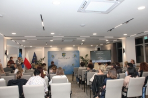 Sala sesyjnej z publicznością siedzącą na krzesłach. Z przodu stoją prowadzący przy mównicy, obok flagi, a w tle widać ekran z prezentacją i planszę Powiatu Cieszyńskiego.