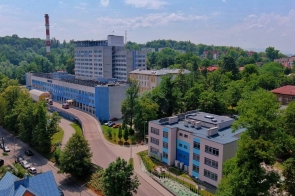 Widok z góry na kompleks Szpitala Śląskiego w Cieszynie. Wśród zieleni znajdują się budynki szpitalne, droga dojazdowa oraz zaparkowane samochody. Tło tworzą drzewa, zabudowania miejskie i jasne niebo.