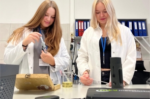 Dwie uczennice w fartuchach laboratoryjnych pracują przy stole w pracowni. Jedna korzysta z mikrometru do pomiaru elementu, druga obsługuje sprzęt pomiarowy przy użyciu sondy. Na blacie znajdują się naczynia laboratoryjne z cieczą oraz aparatura pomiarowa, a w tle widać segregatory z dokumentacją.