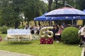 Publiczność siedzi w ogrodzie pod namiotami podczas uroczystości. Na pierwszym planie stoją złote balony z liczbą 65 oraz stolik z białymi torbami upominkowymi.