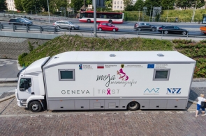 Biały mammobus stoi przy ulicy. Na jego boku widoczne są napisy i logotypy związane z programem „Moja Mammografia”, Funduszami Europejskimi, Unią Europejską, Ministerstwem Zdrowia i NFZ. W tle znajduje się ruchliwa jezdnia z samochodami i autobusem.