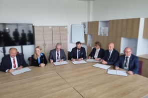 Osoby siedzą przy stole konferencyjnym i podpisują dokumenty. Na blacie widoczne są teczki oraz długopisy, a w tle znajdują się szafy, tablica i ekran.