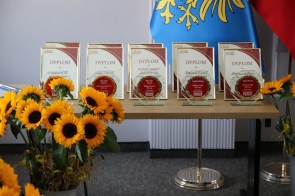 Dyplomy i szklane statuetki ustawione na stole, przygotowane do wręczenia podczas uroczystości. Obok stoją bukiety słoneczników, a w tle widać flagi.
