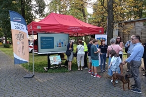 Grupa osób stoi przy czerwonym namiocie Komendy Powiatowej Państwowej Straży Pożarnej w Cieszynie . Pod namiotem znajduje się ekran z prezentacją dotyczącą leków osobistych oraz stół z materiałami informacyjnymi. Obok ustawiono flagę programu OLiOC, a na pierwszym planie stoją uczestnicy.