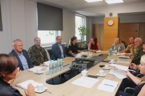 Przy dużym stole w sali konferencyjnej siedzi grupa osób – część w mundurach wojskowych, część w ubraniach cywilnych. Na stole stoją butelki wody, filiżanki z kawą i notatniki, a w centralnym miejscu leży sprzęt multimedialny