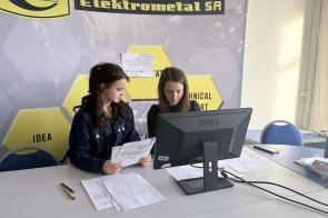 Dwie młode dziewczyny siedzą przy biurku i pracują przy komputerze. Na blacie znajdują się dokumenty oraz telefon, a nad zdjęciem widoczny jest żółty pasek z nazwą firmy Elektometal.