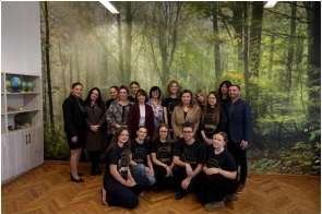 Grupa osób stoi i klęczy do wspólnego zdjęcia w pomieszczeniu z leśną grafiką na ścianie, obok widać regał z globusami.