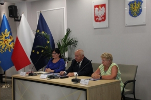 Trzy osoby siedzą przy stole prezydialnym w sali konferencyjnej. Kobieta w niebieskiej sukience, mężczyzna w garniturze i starsza kobieta w pasiastym topie. Za nimi flagi Polski, UE, województwa oraz herby na ścianie.