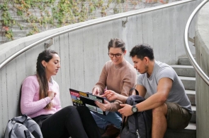 Troje młodych ludzi siedzi na betonowych schodach w otwartym, zielonym otoczeniu. Mają przy sobie plecaki, a jedna z osób trzyma laptop, nad którym wspólnie dyskutują i wskazują coś na ekranie.