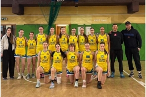 Grupa zawodniczek w żółtych strojach koszykarskich oraz trener na sali gimnastycznej pod koszem do gry.