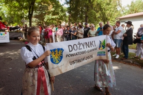 Dwie dziewczynki w strojach regionalnych niosą baner z napisem „Dożynki Powiatowo-Gminne Ustroń 2026”. W tle stoją licznie zgromadzeni uczestnicy i widzowie korowodu.