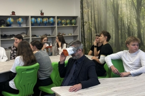 Uczniowie siedzą przy stołach w pracowni z zielonymi krzesłami. Michał Rajwa Naczelnik Wydziału Edukacji korzysta z gogli VR, a w tle widoczne są globusy, minerały oraz fototapeta z motywem lasu.