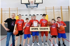 Grupa chłopców w czerwonych koszulkach „Cieszyn” stoi w sali gimnastycznej z medalami, piłkami i dyplomem. Obok nich stoi trener, a z przodu widoczna jest tabliczka „ZPSWR Cieszyn”.