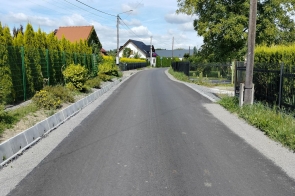 Wyasfaltowana droga biegnie między ogrodzeniami posesji z zielenią i krzewami. W tle widać domy jednorodzinne, a nad nimi niebo z białymi chmurami.