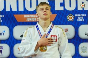 Młody judoka w białym stroju stoi na podium z medalem na szyi, trzymając rękę na piersi. W tle widoczna jest ścianka.