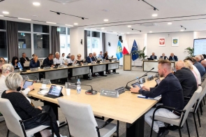 Widok na salę sesyjną podczas obrad. Uczestnicy siedzą przy ustawionych w podkowę stołach, przed nimi znajdują się mikrofony, tablety i tabliczki z nazwiskami. W centralnej części sali widoczne są mównica oraz flagi, a na ścianie godło Polski i herb Powiatu Cieszyńskiego.