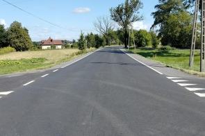 Wyasfaltowana droga biegnie między ogrodzeniami posesji z zielenią i krzewami. W tle widać domy jednorodzinne, a nad nimi niebo z białymi chmurami.