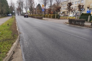 Droga asfaltowa z chodnikiem i znakami drogowymi. W tle widać samochody, drzewa oraz budynek za ogrodzeniem.