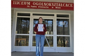 Dziewczyna stoi przed wejściem do liceum i trzyma w rękach dyplom, nad drzwiami widnieje napis z nazwą szkoły w Wiśle.