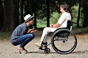Dwie kobiety rozmawiają w parku. Jedna siedzi w wózku inwalidzkim, ubrana na jasno, uśmiecha się. Druga kuca naprzeciw niej, gestykuluje radośnie, nosi kapelusz, jeansy i buty na obcasie. Obie wyglądają na szczęśliwe.