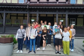 Grupa młodych osób oraz kilka dorosłych stoi na schodach przed drewnianym budynkiem z werandą. Część uczestników trzyma kolorowe plansze lub książki. Wszyscy pozują do wspólnego zdjęcia.