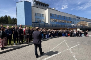 Duża grupa ludzi stoi przed budynkiem i słucha mężczyzny przemawiającego do nich na zewnątrz, w słoneczny dzień przy wejściu głównym.