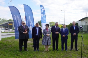 Sześć osób stoi na trawie przy drodze. Obok nich ustawione są niebieskie flagi promocyjne oraz mikrofon na statywie. W tle widać ulicę, samochody i budynki.