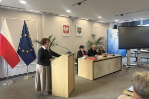 Sala sesyjna, Przy mównicy stoi Janina Żagan Starosta Cieszyński, ubrana elegancko, przemawia do kilku osób siedzących przy stole. W tle flagi i ekran projekcyjny. Na stole mikrofony i dokumenty.