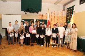 Grupa uczniów i nauczycieli pozuje wspólnie do zdjęcia podczas uroczystości zakończenia roku szkolnego. W tle kolorowa dekoracja z motylkami, sztandar oraz plansze z napisem „Zakończenie roku”.