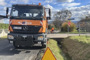 Pomarańczowy pojazd budowlany stoi na remontowanej drodze. Obok widoczne są znaki drogowe, fragment pobocza oraz pracownicy w odblaskowej odzieży w głębi ulicy.
