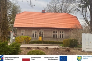 Budynek z nowym czerwonym dachem i odnowioną elewacją, otoczony ogrodzeniem i niską zielenią; przed nim chodnik i krzewy, a u dołu widoczny pasek z logotypami i informacją o dofinansowaniu z funduszy europejskich.