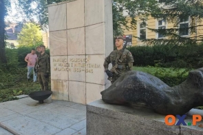 Pomnik z jasną kamienną płytą z napisem oraz ciemną rzeźbą umieszczoną na cokole. Po obu stronach monumentu stoją żołnierze w mundurach, a w tle widoczne są drzewa, krzewy i budynek.
