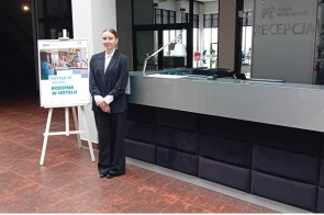  Ewa Raszka w eleganckim stroju hotelowym stoi przy recepcji Term Warmińskich. Obok znajduje się plakat promujący program "Rodzina w hotelu" organizowany przez SGH. Scena oddaje atmosferę profesjonalizmu i gościnności