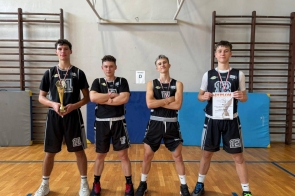 Reprezentacja szkolnej drużyny koszykarskiej po zwycięstwie – czterech zawodników w strojach sportowych prezentuje zdobyty puchar, medale oraz dyplom, stojąc dumnie na sali gimnastycznej po zakończonym turnieju.