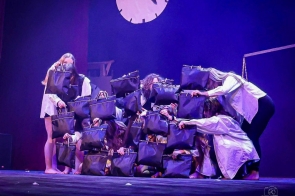 Grupa osób znajduje się na scenie teatralnej oświetlonej fioletowym światłem. Uczestnicy ubrani na biało trzymają czarne torby, którymi częściowo zasłaniają twarze i sylwetki, tworząc zwartą kompozycję.