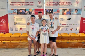 Cztery osoby stoją przed ścianką informacyjną Olimpiad Specjalnych. Trzy osoby z przodu mają białe koszulki z logo wydarzenia, a kobieta pośrodku trzyma dwa medale. W tle widoczne są zdjęcia pływaków.