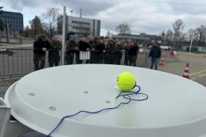 Żółta piłka tenisowa z przywiązanym niebieskim sznurkiem leży na białej antenie satelitarnej. W tle grupa umundurowanych osób stoi za barierką na placu, obok widoczne są pachołki drogowe i biurowce.