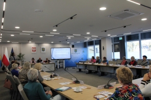 Nowoczesna sala sesyjna z dużymi oknami i jasnym oświetleniem. Przy stołach w kształcie litery „U” siedzą uczestnicy spotkania, słuchając gościa przy mównicy z lewej strony. Na środku stoi ekran z prezentacją, a w tle widoczne są flagi Polski, Unii Europejskiej i województwa śląskiego oraz herby na ścianie.