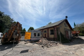 Budynek na terenie prac budowlanych, ogrodzony metalowym płotem. Obok widać koparkę, gruz, materiały budowlane oraz żółtą tablicę informacyjną.