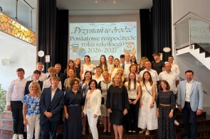 Grupa uczniów i dorosłych pozuje do wspólnego zdjęcia na schodach przed sceną. W tle wyświetlana jest prezentacja dotycząca powiatowego rozpoczęcia roku szkolnego 2026/2027. Po bokach sceny widoczne są zegary oraz kartki z cyframi.