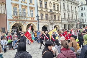 Uczestnicy przechodzą przez rynek z pocztami sztandarowymi. Wokół stoją zgromadzeni mieszkańcy, a w tle widoczne są zabytkowe kamienice.