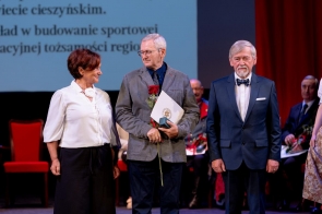 Trzy osoby stoją na scenie podczas uroczystości. Mężczyzna pośrodku trzyma różę, dokument oraz małe pudełko, a w tle widoczne są czerwone kurtyny i ekran z tekstem.